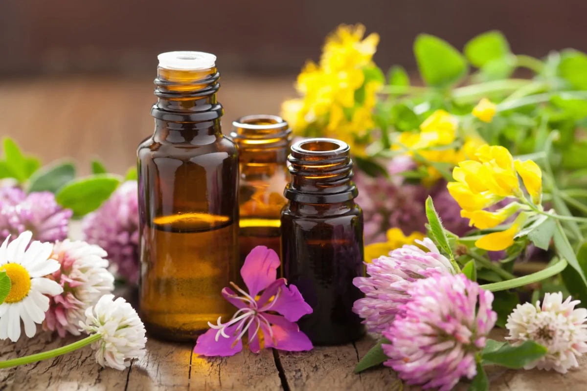Three small amber glass bottles surrounded by colorful flowers, including daisies, clover, and other wildflowers, placed on a rustic wooden surface.