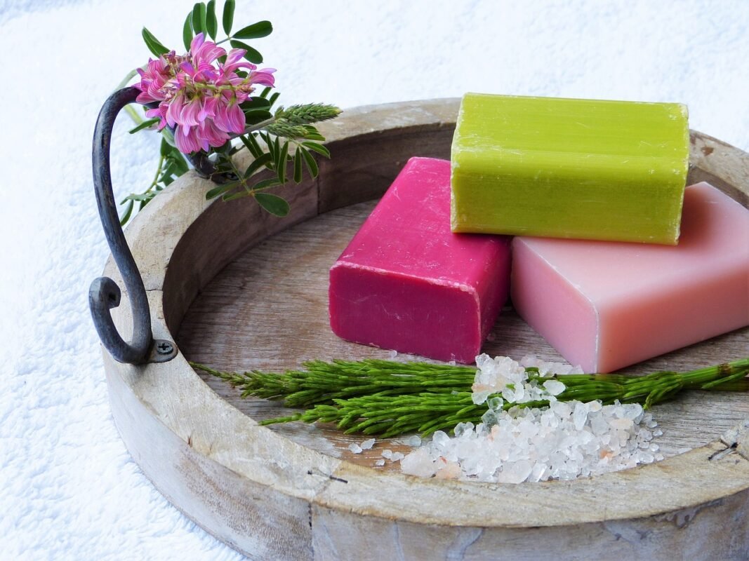 Three Rectangular Soap Bars in Green, Pink, and Light Pink, Arranged on a Wooden Tray with Metal Handle, Accompanied by Pink Flowers, Green Sprigs, and Coarse White Salt Crystals.