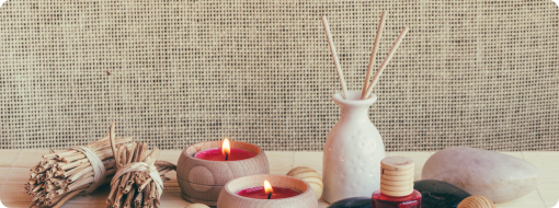 Two lit candles in wooden holders, white vase with reed diffusers, dried herb bundle, essential oil bottle, and smooth stone on a burlap background.