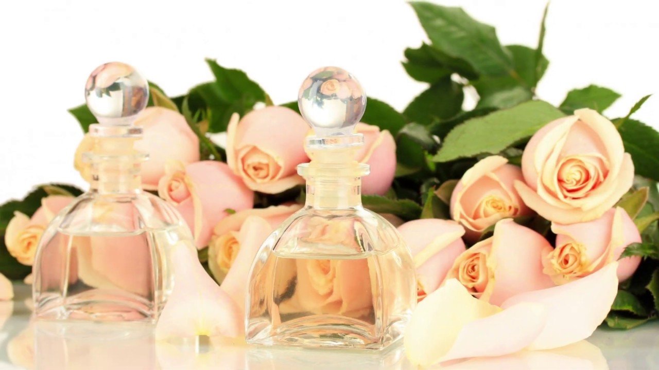 Two glass perfume bottles with spherical stoppers, placed in front of a bouquet of light pink roses with green leaves, and scattered rose petals around them.